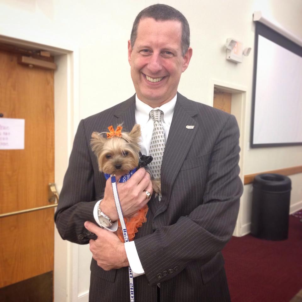 Dr. Reuban Rodriguez of V C U is wearing a gray suit and smiling while holding Dogs On Call therapy dog Winnie, a yorkie.